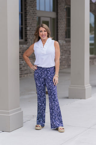 A woman stands confidently in front of a modern boutique pillar wearing the Midnight Maritime Pants, featuring a dramatic navy and white botanical leaf print. These high-waisted wide-leg trousers offer a structured yet flowing silhouette and are paired with a white ruffled-collar sleeveless polo and tan woven platform slides.