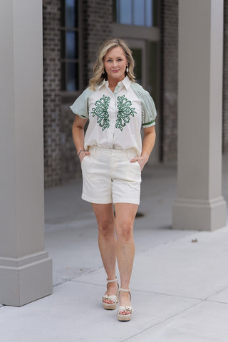 A woman stands in front of a boutique wearing the Emerald Estate Button-Down Top, a sophisticated cream blouse featuring intricate emerald green scrolling embroidery on the chest. The top is designed with olive-striped 1/2 length puff sleeves and is styled with ivory shorts and cream platform sandals for a polished, "old money" spring look.