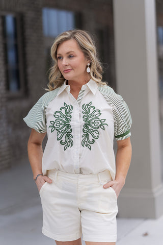 A detailed portrait view highlighting the breathtaking symmetrical emerald green embroidery on the Emerald Estate Button-Down Top. The high-contrast design features a structured pointed collar and 1/2 length puff sleeves with olive green vertical stripes and solid green ribbed cuffs.