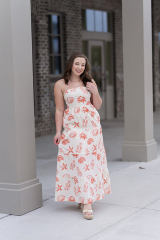 A woman stands in front of a boutique pillar wearing the High-Tide Tiered Dress, a strapless maxi-length silhouette featuring a flowing tiered skirt. The dress showcases a whimsical red nautical print of lobsters, crabs, starfish, and seashells on a crisp cream background. She is styled with cream platform sandals and a gold link necklace for a polished, "resort-ready" aesthetic.