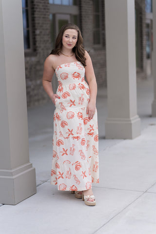 A woman stands in front of a boutique pillar wearing the High-Tide Tiered Dress, a strapless maxi-length silhouette featuring a flowing tiered skirt. The dress showcases a whimsical red nautical print of lobsters, crabs, starfish, and seashells on a crisp cream background. She is styled with cream platform sandals and a gold link necklace for a polished, "resort-ready" aesthetic.