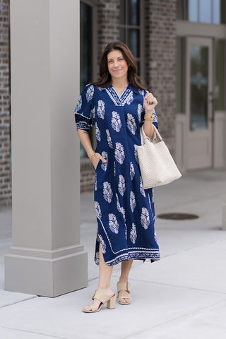 A woman stands in front of a modern boutique wearing the Ocean Odyssey Midi Dress, a deep navy boarder print collared midi dress. The dress features a sophisticated split V-neckline, half-length puff sleeves with patterned trim, and an intricate ivory botanical crest print organized in a striking border layout. She has styled it with tan block-heel mules and a large white woven tote bag for a "resort-chic" aesthetic.