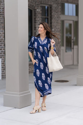 A woman stands in front of a modern boutique wearing the Ocean Odyssey Midi Dress, a deep navy boarder print collared midi dress. The dress features a sophisticated split V-neckline, half-length puff sleeves with patterned trim, and an intricate ivory botanical crest print organized in a striking border layout. She has styled it with tan block-heel mules and a large white woven tote bag for a "resort-chic" aesthetic.