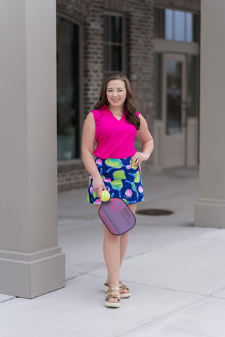 A woman stands in front of a modern boutique pillar wearing the Pink Punch Polo, a vibrant sleeveless top in "Hot Pink". The polo features a feminine V-neck and a structured collar. She has styled it for a high-energy athletic look with the Pickleball Pro Skort, featuring a navy base with neon green and pink motifs, and gold metallic platform sandals.