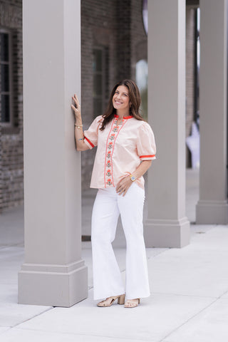 A woman stands in front of a modern boutique pillar wearing the Palm Beach Patio Top, a sophisticated short-sleeve blouse in a warm sandy beige. The top features a romantic red ruffled stand-up collar and vibrant vertical floral embroidery in coral-orange and green cascading down the center. She has styled it with high-waisted white flare trousers and tan block-heel sandals for a polished, "preppy-chic" summer look.