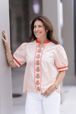 A detailed portrait view of the Palm Beach Patio Top highlighting the intricate, symmetrical floral embroidery and the feminine notched V-neckline. The elegant puff sleeves are shown with matching red contrast bands at the cuffs, creating a structured yet soft shoulder line. The model is accessorized with gold stacked bracelets and a gold watch to complement the tropical luxury aesthetic.