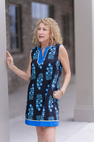 A portrait view of the Midnight Meadow Mini highlighting the intricate botanical print and the sophisticated notched V-neckline. The high-contrast blue border and lime green piping frame the face, while the model demonstrates the functional discreet side-seam pockets. The premium woven fabric offers a crisp, tailored look perfect for elevated daytime events or garden parties.