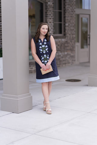 A woman stands in front of a modern boutique pillar wearing the Midnight Meadow Mini Dress, a sleeveless A-line mini dress in deep navy. The dress features a sophisticated notched V-neckline and intricate, symmetrical floral embroidery in sky blue and sage tones cascading down the center. She is styled with a woven raffia clutch and cream-colored platform sandals for a sophisticated resort aesthetic.