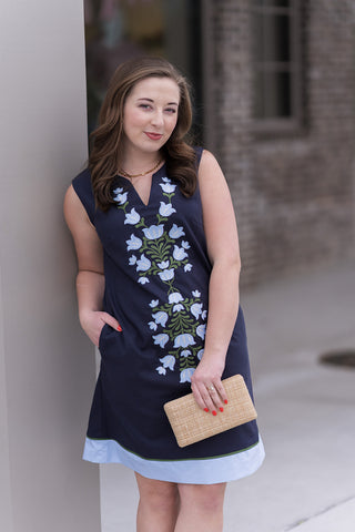 A detailed portrait view of the Midnight Meadow Mini Dress highlighting the breathtaking wildflower-inspired embroidery. The high-density thread-work in sky blue and sage green creates a vertical, elongating effect against the rich nocturnal navy canvas. The model is accessorized with a gold link necklace to complement the elegant notched neckline.