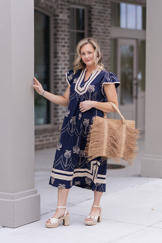 Front view of the Sandy Shores Midi Dress highlighting the intricate geometric motifs along the hemline. The model is carrying a large fringed straw tote bag and wearing white drop earrings, illustrating a tropical luxury aesthetic perfect for an upscale seaside dinner.
