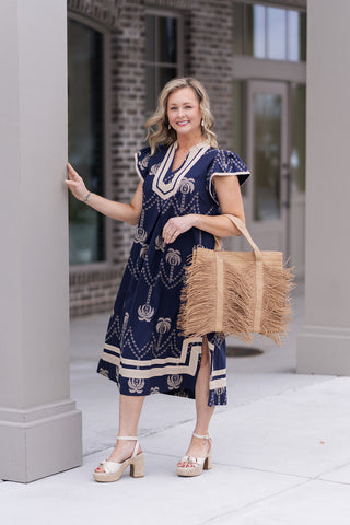 Front view of the Sandy Shores Midi Dress highlighting the intricate geometric motifs along the hemline. The model is carrying a large fringed straw tote bag and wearing white drop earrings, illustrating a tropical luxury aesthetic perfect for an upscale seaside dinner.