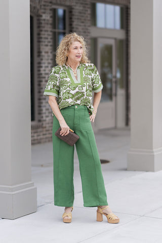 A woman stands in a boutique setting wearing the Palm Pavilion Top, a vibrant green-and-white botanical top featuring a tropical palm and tree motif. The top is designed with a notched V-neckline framed by a bold green border and half-length puff sleeves with striped wide-banded cuffs. She has styled it for a "resort-chic" aesthetic with the high-waisted, wide-leg Clubhouse Classic Pants in a rich fern green, featuring prominent front patch pockets and tan woven platform heels.