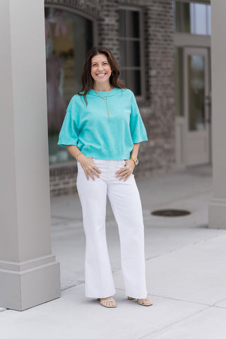A woman stands in front of a modern boutique wearing the Seafoam Serenity Knit Top, a vibrant seafoam green pullover in a premium lightweight sweater knit. The top features a modern drop-shoulder design and is styled for a sophisticated "coastal chic" look with high-waisted white flare trousers and a gold y-neck chain necklace.