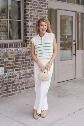A woman stands in front of a modern boutique wearing the Scalloped Shoreline Wide-Leg Pants, a pair of crisp white high-waisted trousers. The pants feature a flattering wide-leg silhouette and are styled with a white sleeveless knit vest with green horizontal stripes, tan woven platform slides, and a small woven clutch for a "clean girl" maritime look.