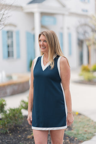 A detailed profile view of the Game, Set, Match Mini Dress highlighting the premium navy knit fabric and the clean white binding at the V-neckline. The photo emphasizes the athletic-inspired silhouette and the model’s accessorized look with a gold pendant necklace and delicate beaded bracelets.