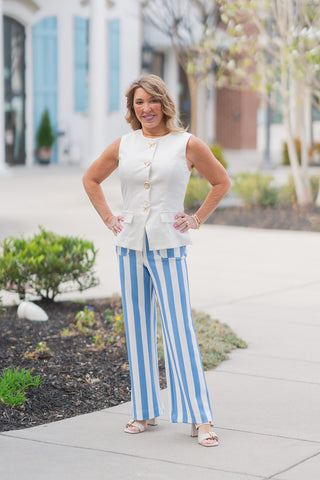 Model posing in the Gilded Reef Vest, a structured white sleeveless tailored top featuring ornate golden marine-themed buttons. She is styled with high-waisted blue and white vertical striped wide-leg trousers and cream block-heel sandals. The outfit is set against a bright, upscale outdoor plaza, capturing a "nautical chic" resort aesthetic.