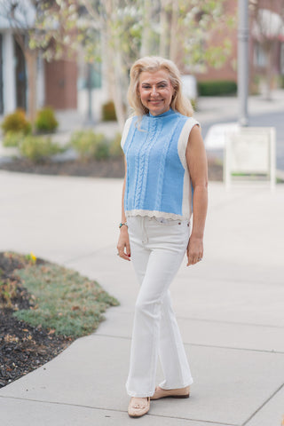 A full-length front view of a woman wearing the Azure Horizon Vest, a sophisticated medium-weight knit vest in a serene light blue and cream color-block design. The vest features an intricate vertical cable knit pattern on the vibrant blue center panel and a polished high mock-neck collar. It is styled for a "coastal chic" look with high-waisted white denim and tan loafers, set against a bright, upscale outdoor backdrop.