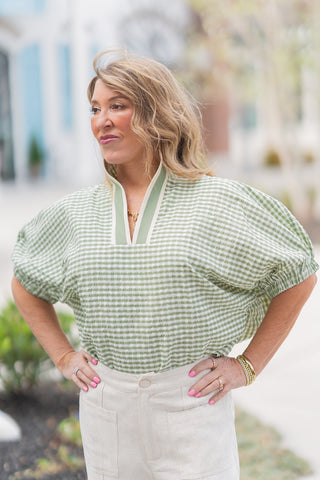 A detailed front view of the Garden Gingham Top in Matcha green, highlighting the architectural contrast trim along the V-neckline. The shot emphasizes the lightweight woven fabric and the subtle elasticated cuffs that create a gentle puff effect on the dolman sleeves. The model is accessorized with a gold link necklace and stacked gold bracelets.