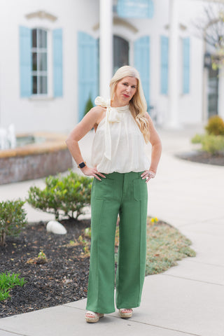 Model posing in the Ivory Illusion Bubble Top, a crisp ivory halter-style top featuring a voluminous bubble hem and an elegant self-tie neck detail. She is styled with high-waisted green wide-leg trousers and cream platform sandals. The top's structured pleating and breezy silhouette create a modern boutique aesthetic against a bright, Mediterranean-inspired outdoor backdrop.