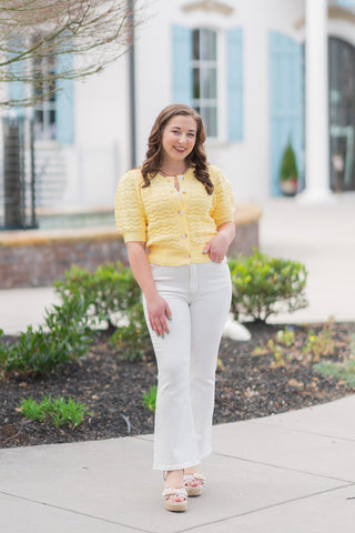 Model posing in the Daffodil Dream Cardigan, a vibrant buttery yellow short-sleeve knit featuring an intricate zigzag open-knit pattern. The cardigan is styled fully buttoned as a top, paired with high-waisted white flare jeans and cream platform espadrille sandals. The look is set outdoors against a bright, Mediterranean-style architectural background, capturing a fresh spring "boutique chic" aesthetic.