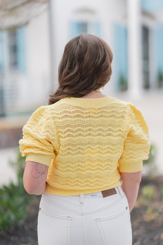 Back view of the Daffodil Dream Cardigan emphasizing the consistent allover texture and the structured silhouette of the puff sleeves. The wide rib-knit hem sits perfectly at the waist of white "Judy Blue" branded denim, demonstrating a flattering, cinched fit. This view highlights the high-quality construction and the rich, multidimensional look of the yellow artisanal crochet-style knit.