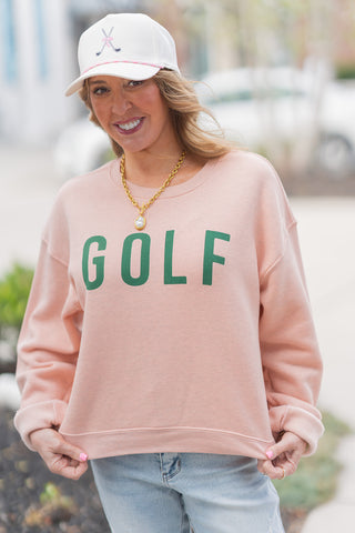 A portrait-style close-up highlighting the "quiet luxury" details of the Country Club Pullover. The shot emphasizes the vibrant green contrast of the "GOLF" lettering against the premium, heathered pink sponge fleece fabric. The model is accessorized with a gold chain necklace featuring a large pearl pendant and a white baseball cap with a pink golf club logo, showcasing a sophisticated, sporty-chic aesthetic.