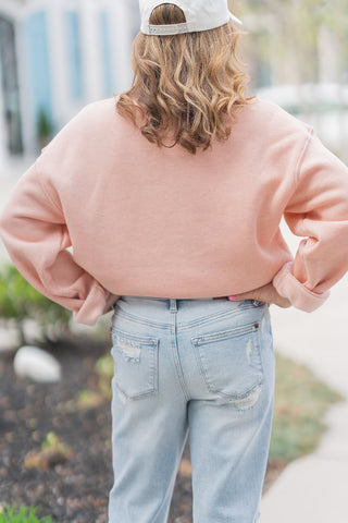 A full-length back view of the Country Club Pullover showcasing its clean, streamlined profile and high-quality construction. The heathered pink fabric provides a soft, feminine base, highlighting the extra-long sleeves and the sturdy ribbed waistband that sits perfectly at the natural waistline. Paired with distressed denim and a white cap, this angle demonstrates the pullover's effortless "cool-girl" confidence.