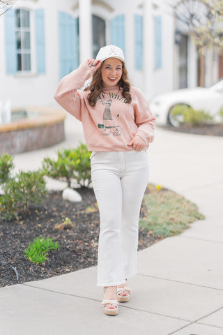A full-length front view of a woman posing in the Golf Wives Social Club Pullover, a soft heathered pink sweatshirt featuring a vintage-inspired forest green graphic of a female golfer. The pullover is designed with a comfortable boxy fit and a trendy mid-length hem. It is styled for a "clubhouse chic" look with high-waisted white flared denim, a matching "Golf Wives" baseball cap, and cream platform sandals. The model is standing in a bright outdoor plaza with light blue shutters in the background.