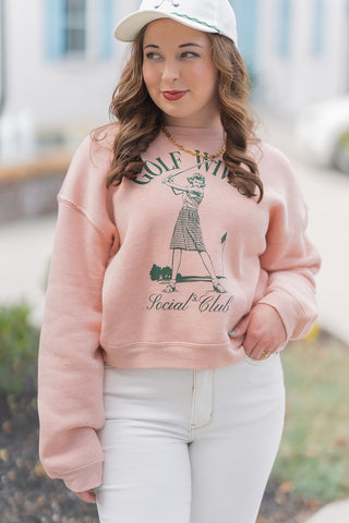A portrait-style close-up highlighting the retro-inspired details of the Golf Wives Social Club Pullover. The shot showcases the deep forest green "Golf Wives Social Club" script and the detailed illustration of a golfer in mid-swing. The ultra-plush sponge fleece fabric and classic crew neck are visible, along with the model's gold chain necklace and coordinating baseball cap, emphasizing a sophisticated yet sporty boutique aesthetic.