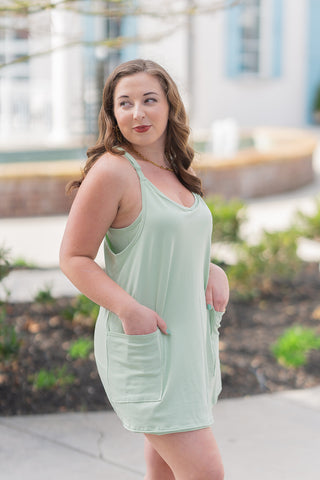 A profile shot of a woman wearing the sage green Pocket-Full-Of-Sunshine Romper, emphasizing the deep side patch pockets and the comfortable, relaxed fit. She is looking over her shoulder, styled with a gold chain necklace and white athletic sneakers for a sporty, functional summer look.