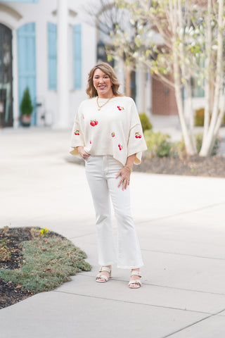A woman posing outdoors in Judy Blue Cloud Walk Jeans, which are high-waisted white kick flare denim pants. She is wearing them with an oversized cream-colored knit poncho sweater adorned with embroidered red cherries, nude block-heeled sandals, and gold jewelry. The jeans feature a body-hugging fit that transitions into a subtle flare at the calf, hitting just above the ankle.