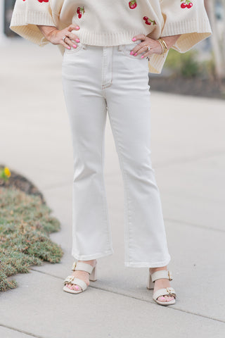 A close-up front view of the Judy Blue Cloud Walk Jeans, highlighting the pristine white wash and high-quality opaque fabric. The detail showcases the high-waisted design with a traditional button and zip-fly closure, functional front pockets, and subtle tan-toned contrast stitching. The slim fit through the thighs flares out at the hem, perfectly complementing the buckled cream sandals.