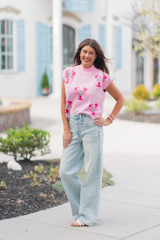 A woman stands smiling in a pair of light-wash Judy Blue wide-leg jeans, featuring a unique "v-shape" western-style yoke at the waistband and a relaxed, floor-length silhouette. She has styled the denim with a sleeveless baby pink knit mock-neck sweater adorned with vibrant hot pink and green lobster motifs. The look is completed with white embellished slide sandals, posed on a clean outdoor walkway with a blurred coastal-style building in the background.