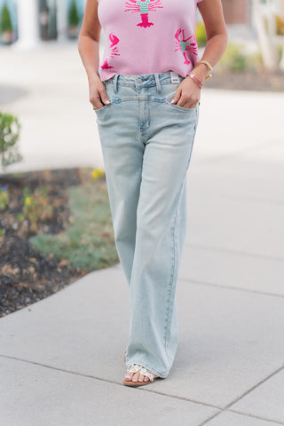 A close-up front view of light-wash wide-leg jeans highlighting the decorative stitching on the high-rise waistband and the functional front pockets. The denim features a subtle vintage fade and a voluminous, straight-fall leg that skims the floor. The subject’s hands are tucked into the pockets, showcasing the soft drape of the pink lobster-print knit tucked into the seamless waist.