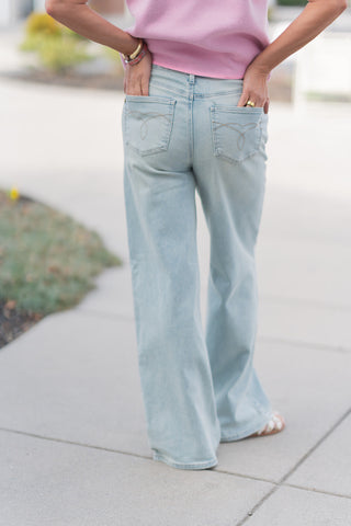 The back view of the light-wash Judy Blue jeans, showcasing the signature rear pocket embroidery with a minimalist looped "infinity" design in tan contrast stitching. The high-waisted fit contours the figure before dropping into a wide, breezy leg. The smooth, lightweight denim finish and clean hemline are visible, emphasizing the premium construction and "lived-in" comfort of the fabric.