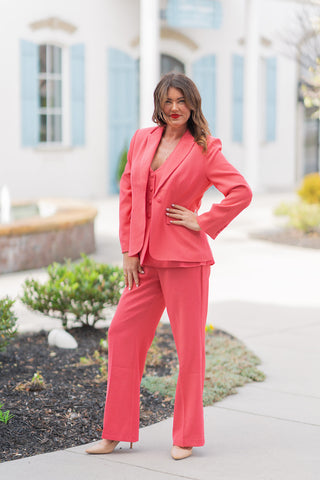 A full-length view of a woman posing in the Watermelon Sugar Blazer, a vibrant coral-pink power blazer. The model wears a sophisticated three-piece suit, pairing the structured, single-breasted blazer with a matching tonal vest and high-waisted wide-leg trousers. The blazer features sharp notched lapels, tonal fabric-covered buttons, and functional flap pockets. Styled with nude pointed-toe pumps against a bright, upscale outdoor backdrop with light blue shutters.