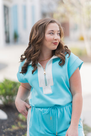 Close-up views of the Reef Runner Top highlighting the functional gold-toned zip-front closure framed by a crisp white contrast placket. The top features athletic cap sleeves and a tailored collar, crafted from a premium, non-sheer lightweight knit fabric. The vibrant turquoise and white palette provides a refreshing, high-energy seasonal aesthetic.