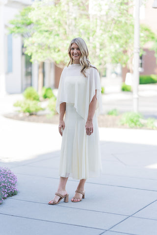 A woman stands outdoors smiling in the Sunbeam Silhouette Midi Dress, a two-piece ensemble in a soft primrose yellow. The outfit features a structured slip-style midi dress with a sheer, ethereal capelet overlay that drapes elegantly over the shoulders. She has styled the look with tan braided block-heel sandals, creating a sophisticated and airy aesthetic perfect for a summer gala or garden party.