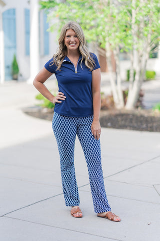 A woman stands smiling in a sunny outdoor setting wearing the Riviera Rope Pants, a pair of "Charlene" style wide-leg trousers. The pants feature a vibrant navy and cream geometric honeycomb lattice pattern. She has styled them with a coordinating navy short-sleeve quarter-zip polo and tan leather slide sandals, creating a polished, nautical-inspired resort look.