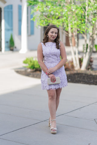 A woman stands outdoors wearing the Lavender Haze Petal Mini Dress, a stunning shift dress featuring all-over 3D floral appliqués on delicate lavender lace. She has styled the look with cream-colored platform espadrille heels and a textured beige clutch. The dress hits at a flirty mini length with a sheer tulle hem, creating an ethereal, romantic aesthetic perfect for a spring wedding or garden party.