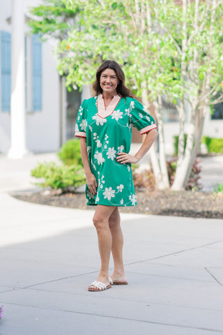 A woman stands outdoors wearing the Wildwood Vine Mini Dress, a vibrant emerald green shift dress featuring all-over white floral vine embroidery. The dress is styled with cream-colored embellished slide sandals. The silhouette is a relaxed mini-length fit, showcasing the bold contrast between the lush green fabric and the delicate botanical needlework against a bright, suburban background.
