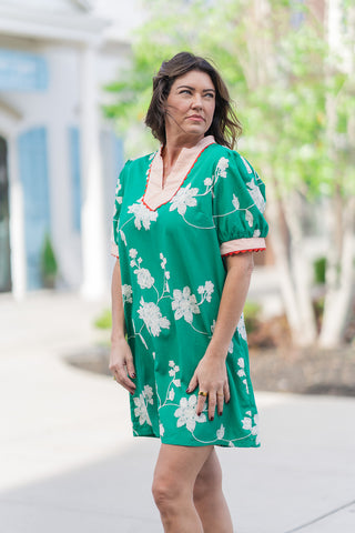 A three-quarter view of the Wildwood Vine Mini Dress highlighting the intricate neckline. The split V-neck is trimmed with a peach-and-white striped contrast fabric and a tiny red scalloped edge. The short puff sleeves feature matching striped cuffs and red trim, adding a playful, artisanal touch to the structured emerald woven fabric and white floral embroidery.