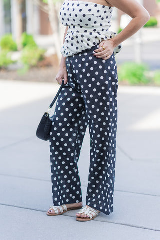 A detailed front-angle view of the Mon Chéri Dotted Pants highlighting the flowy wide-leg silhouette and high-quality woven fabric. The inverted polka dot pattern creates a striking visual contrast, while the high-rise waistband provides a structured, elongating fit. The image showcases the dramatic volume and effortless drape of the pants as they graze the top of the woman's sandals.