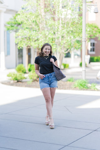 A woman walks outdoors wearing the Z Supply Modern Slub Tee in black, tucked into high-waisted Vervet denim shorts with a raw hem. She styles the classic crew neck tee with platform espandrill sandals, a gold pearl necklace, and a large sparkling tote bag. The outfit represents a perfect casual-chic spring transition look.
