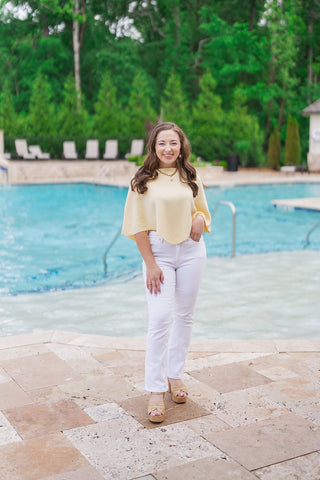 A full-length photograph of a woman wearing the Bellini Breeze Top, standing on a travertine patio by a swimming pool with a waterfall and lush green trees. The fine-gauge, "Butter Yellow" knit top features a draped boat neckline and a unique handkerchief pointed hemline. It is styled with crisp white straight-leg pants and natural fiber platform sandals, creating an elegant and breezy coastal aesthetic.