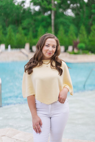 A portrait view detailing the Bellini Breeze Top, showcasing the delicate, premium fine-gauge knit texture and the sophisticated slouchy boat neckline. The unique poncho-style drape frames the model’s torso and arm. She has long wavy hair, is looking directly at the camera, and wears a gold chain necklace with a pearl pendant, emphasizing the top as a refined "quiet luxury" piece.