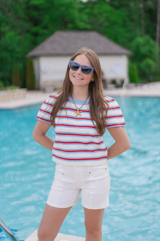 A woman stands poolside wearing the Dear John Julian White Shorts, a pair of polished optic white denim shorts with a mid-high rise and clean-hem finish. The shorts feature unique vertical front seams that elongate the silhouette. She has styled them with a red, blue, and white striped knit top, blue sunglasses, and a gold starfish necklace for a nautical, preppy spring look.