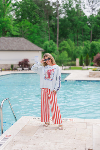 A full-length view of a woman standing poolside wearing the Salty Stamp Pullover, a pale "Sea Salt Blue" sweatshirt featuring a large nautical postage stamp graphic with a red lobster and navy scalloped borders. She is styled with high-waisted red and white vertical striped wide-leg trousers, cream-colored block heel sandals, and blue-rimmed sunglasses, creating a vibrant, patriotic-inspired coastal aesthetic.