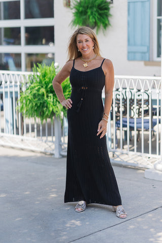 Woman smiling and posing outdoors in the Z Supply Sunda Midi Dress, a black crochet knit maxi dress featuring a scoop neckline and thin adjustable camisole straps. The dress is designed with a small keyhole cutout below the bust, accented by a functional drawstring with wooden beads and tassel ends. The style is completed with a thick gold chain statement necklace with a coin pendant, stacked gold bracelets, and metallic silver low-block heeled sandals.