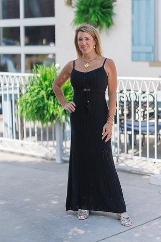 Woman smiling and posing outdoors in the Z Supply Sunda Midi Dress, a black crochet knit maxi dress featuring a scoop neckline and thin adjustable camisole straps. The dress is designed with a small keyhole cutout below the bust, accented by a functional drawstring with wooden beads and tassel ends. The style is completed with a thick gold chain statement necklace with a coin pendant, stacked gold bracelets, and metallic silver low-block heeled sandals.